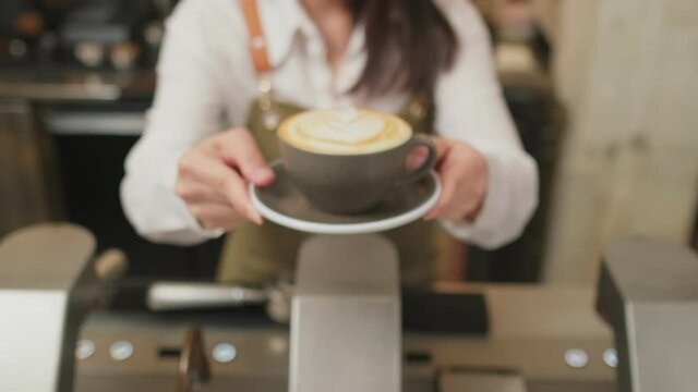 Asian female barista holding artwork latte coffee serving to customer.