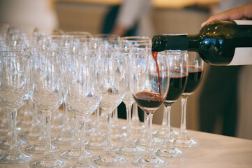 hand of a waiter do pouring red wine from bottle to wineglass that set up for dinner reception party in evening time.
