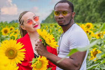 A young couple in love, a guy and a girl, stand in the middle of a field of sunflowers and look at the camera