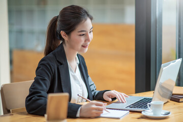 Charming asian businesswoman working on laptop and taking note while sitting at the table in office.
