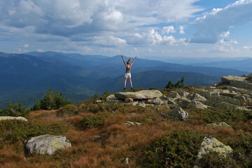 A woman jumps, dances, and climbs, does yoga on top of mountains. Young woman hiking up hill against a blue sky with clouds.