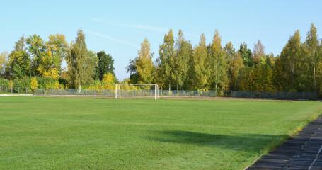 In an empty stadium, a green football field and white gates against the backdrop of an autumn forest and blue sky.