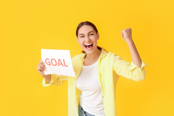 Happy young woman holding paper with word GOAL on color background