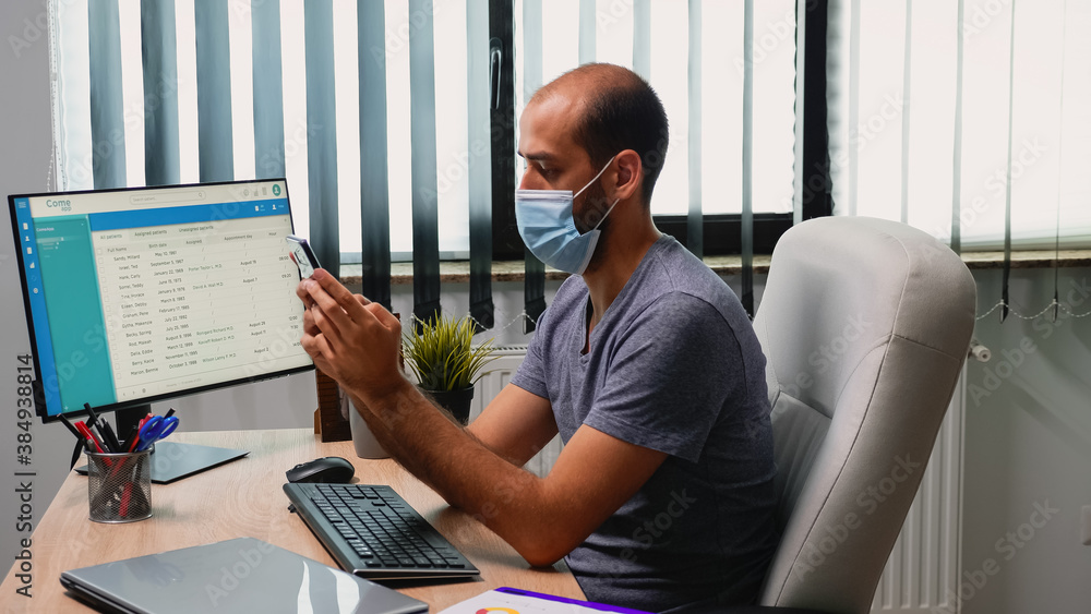 Poster man sitting in office room on desk typing on phone wearing protection mask. freelancer working in ne