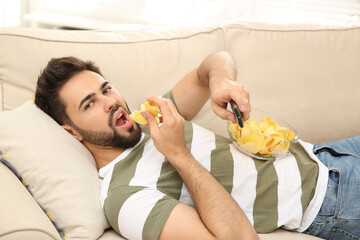 Lazy young man with potato chips watching TV on sofa at home