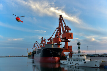 In the sunset, the crane is lifting goods from the wharf to the transport ship. And there are planes flying by.