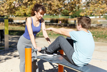 Tweenage boy doing push-ups on sport bench while mother controlling process of training