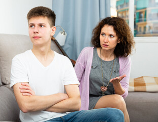 Portrait of troubled teen boy and his mother scolding him in home interior..