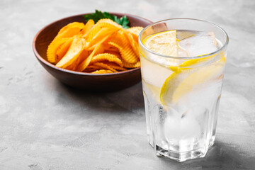 Fresh ice cold water drink with lemon near to fried corrugated golden potato chips with parsley leaf in wooden bowl on concrete background, angle view