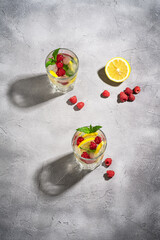 Fresh cold ice water drink with lemon, raspberry fruits and mint leaf in two faceted glass on stone concrete background, summer diet beverage, angle view