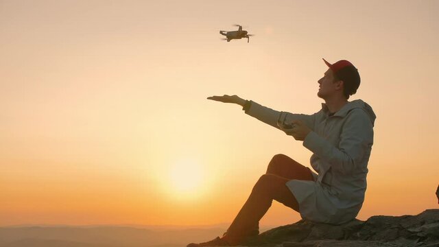 Man sitting on edge of rock cliff and starts drone from his hand at sunset