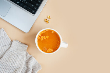 hot soup with crackers in mug, omega-3 vitamins, silver laptop and cozy knitted beige sweater on pastel beige table. remote work from home during diseases period, top view, selective focus