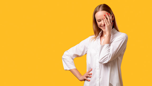 Shy woman. Fun joke. Positive attitude humor. Female lifestyle. Amused lady in white shirt smiling with facepalm gesture isolated on orange empty space commercial background.