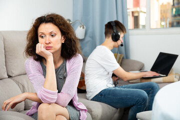 Obraz premium Frustrated mother sitting on sofa at home while her teenage son absorbed of social networks on laptop