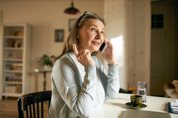 Modern gray haired woman pensioner holding cell phone and raising fore finger, ordering food using delivery service. Mature female speaking to friend using electronic gadget, sitting at home
