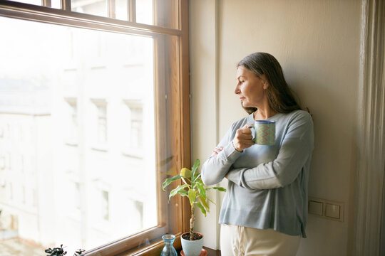 Social Distancing, Aging And Lifestyle Concept. Portrait Of Thoughtful Gray Haired Senior Woman Staying Home Standing By Window Holding Mug, Drinking Morning Coffee, Looking Outside With Interest