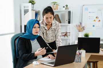 Obraz na płótnie Canvas Casual pregnant businesswoman in suit and headscarf working with colleague in office. young maternity muslim lady worker using laptop computer at desk and talking to coworker. teamwork solve problem.