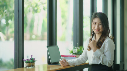 Obraz premium Portrait of a young attractive Asian woman relaxing while sitting at her office desk, smiling and looking at the camera.