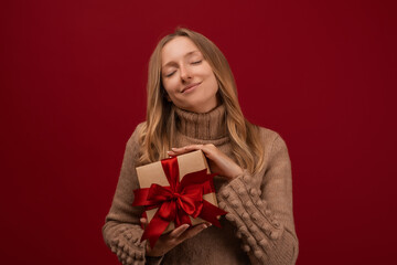 Charming blonde woman holding gift with red ribbon. Studio shot red background. New Year Birthday Holiday concept