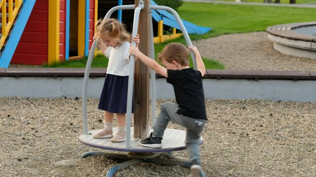 Adorable little boy and girl ride merry-go-round spending time on large contemporary playground in sunny summer city park