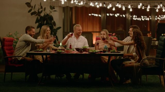 Friends sitting at dining table on backyard of house. Positive man making toast