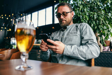 Middle aged bearded man sitting in bar and using smart phone for hanging on social media sites. In foreground is glass of cold fresh beer.