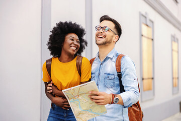 Young attractive multicultural laughing couple walking outdoors and visiting city. Man having map in his hands. Tourism concept.