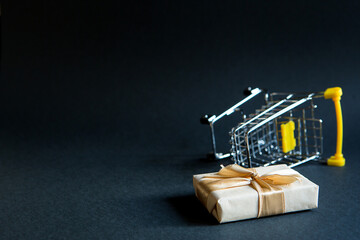 Fototapeta premium Box with a gift in Christmas packaging in a shopping cart on a black background. Black Friday, buying gifts for the new year. Space for text