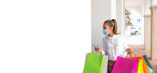 Beautiful fashionable blonde woman doing shopping holding colored shopping bags, wearing a medical mask, looking to a big white copy space with a smartphone in her hand - Female Coronavirus shopper