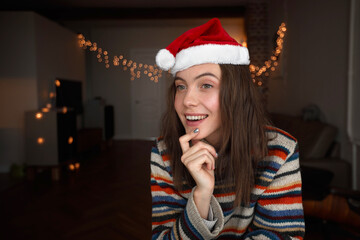 Happy dreamy young female in striped knitted sweater and red Christmas hat sitting in dark room with glowing garlands