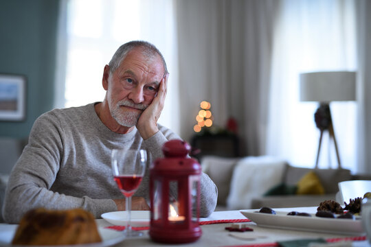 Lonely senior man with wine sitting at the table indoors at Christmas, solitude concept.
