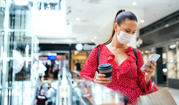 Woman with face mask standing and using smartphone indoors in shopping center, coronavirus concept.