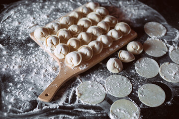 homemade dumplings lie on a wooden cutting board on the kitchen table, flour and dough circles are scattered nearby. Image with selective focus and noise effect