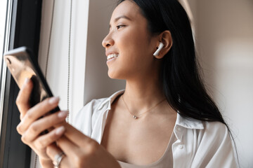 Charming cheerful asian woman in earphones using mobile phone