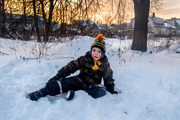 Fototapeta na wymiar The kid in overalls is sitting in the snow and smiling. Portrait of a child in the snow.
