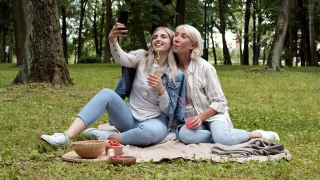 Wide shot of young pretty Caucasian woman and her middle-aged mom sitting together on plaid outdoors, having picnic and making selfie using phone