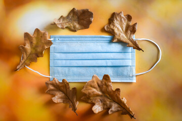A medical protective mask and yellow dry oak leaves lie on a bright autumn background. The concept...