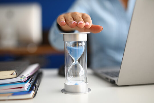 Woman holds her hand over hourglass at workplace in office closeup. Time is money concept.