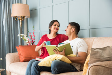 Asian Indian smart couple reading books while sitting on sofa at home