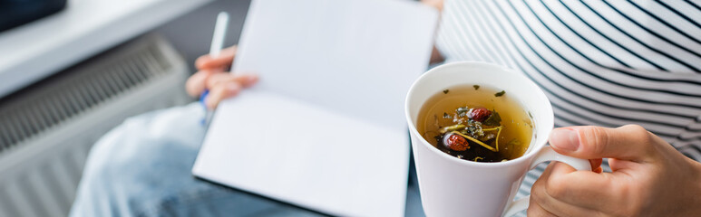 Panoramic shot of woman holding cup of herbal tea and notebook at home
