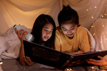 Family concept. elder sister and sister reading book with flashlight together in children tent...