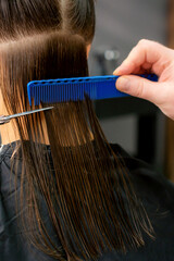 Close up of hands of male hairdresser cutting long hair of young woman holding scissors and comb at the salon. Rear view