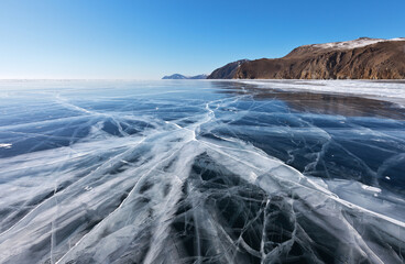 Beautiful winter landscape of frozen Baikal Lake with transparent smooth ice with cracks near the rocky shore in sunny cold day. Winter ice travel concept. Natural background