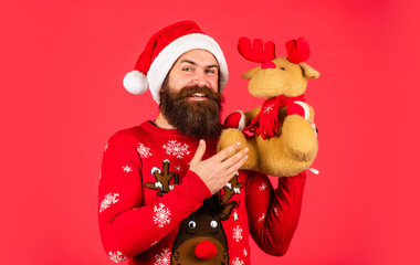 brutal man hold reindeer toy. christmas online shopping. bearded hipster in xmas sweater. winter holiday party. mature guy celebrate new year. man with beard in santa claus hat