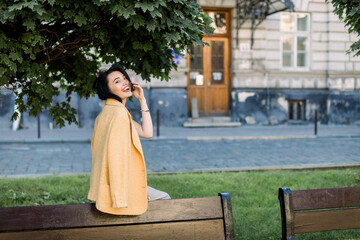 Laughing elegant Asian woman in yellow jacket, chilling and having fun during her morning city walk. Outdoor photo of lovely brunette business lady sitting on bench.