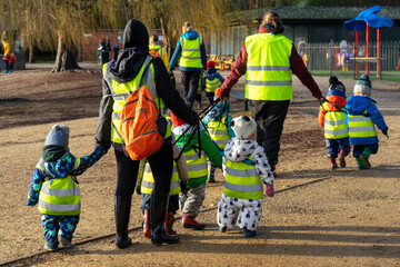 Nursery age children walking together with preschool teachers through a park wearing high visibility jackets