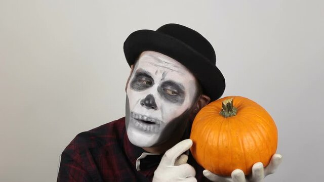  A scary clown with a pumpkin on his head grimaces and laughs terribly.   A terrible man in clown makeup and with a pumpkin on his head grimaces and waves his hand in greeting to his victim.