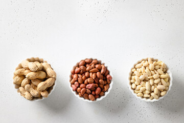 Peanut in three types with peel, roasted and peeled in bowls on white table with copy space.