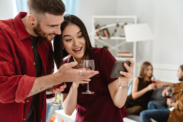 Image of joyful multinational friends using cellphone while drinking wine