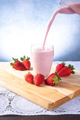 A pitcher pouring strawberry juice into a glass on wood with strawberries around it, selective focus.